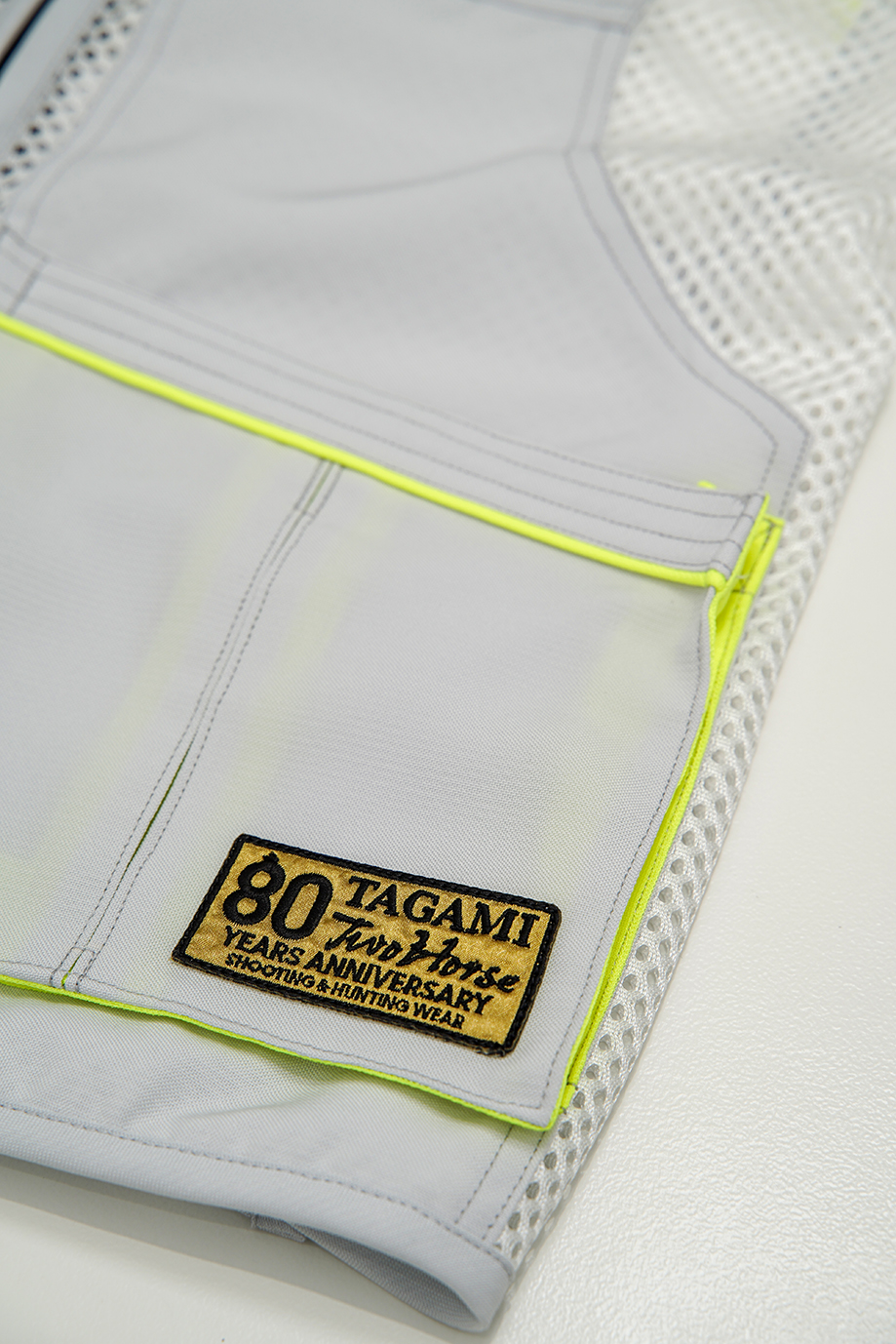 DSC08284_low – f-range 東京表参道の銃砲店 Close-up of light gray fabric with neon yellow zipper trim and a rectangular patch reading '80 TAGAMI Years Anniversary Shooting & Hunting Wear' on a jacket leg patch pocket.